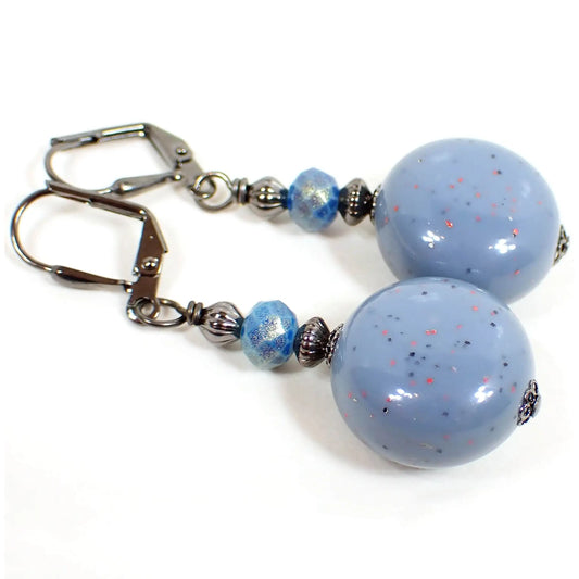 Angled view of the handmade drop earrings. The metal is gunmetal gray in color. There are faceted blue glass beads at the top that have a golden sheen on them. The bottom beads are reclaimed vintage confetti lucite beads and are a country blue in color with tiny flecks of blue and pink confetti glitter embedded in them. They are a puffy round shape.