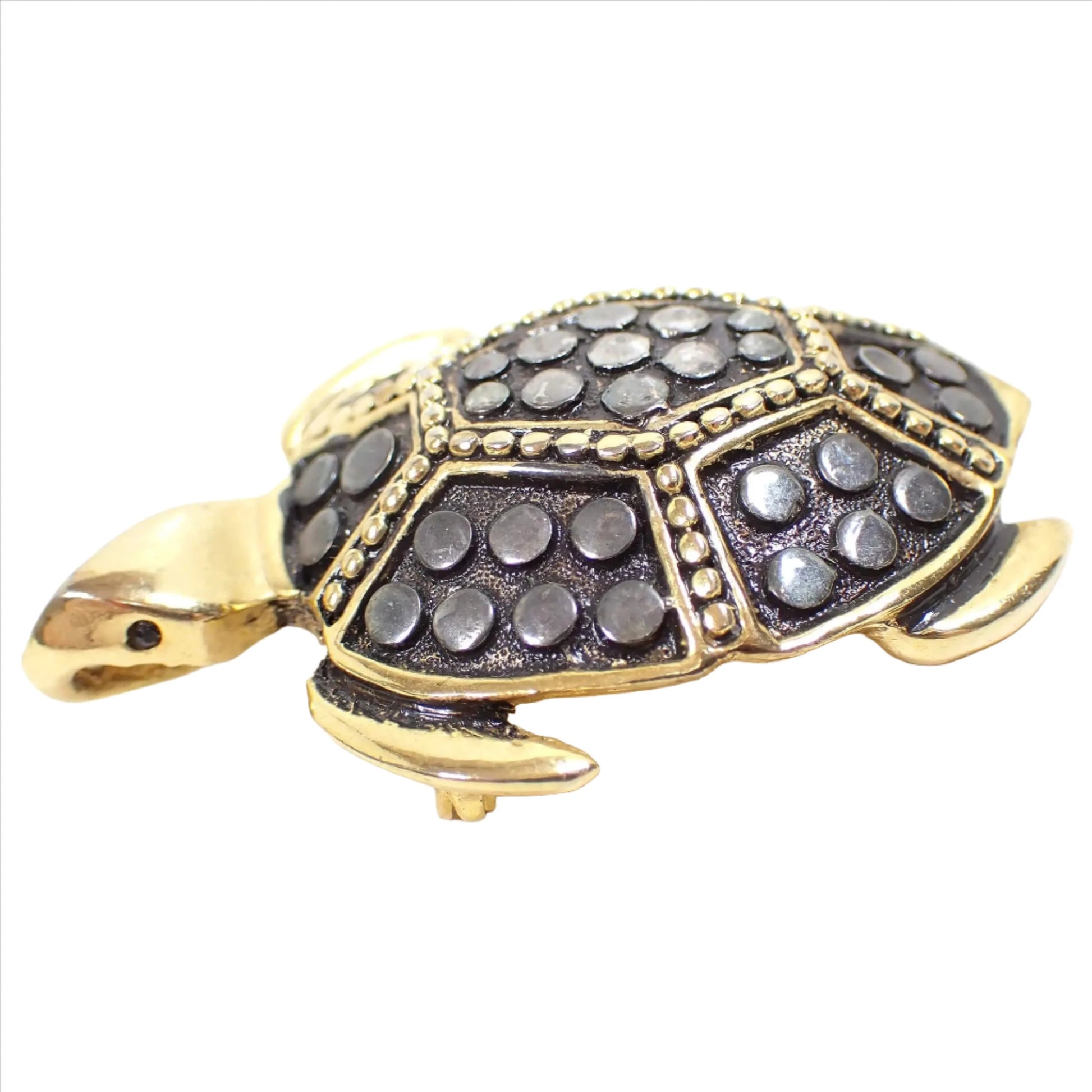Angled side view of the retro vintage SY pendant brooch pin. The brooch is shaped like a turtle that is gold tone in color with gunmetal color areas on the shell. There are dark gray faceted eyes and the part under the mouth is where the pendant bail is built in if you want to put it on one of your chains to wear as a pendant.