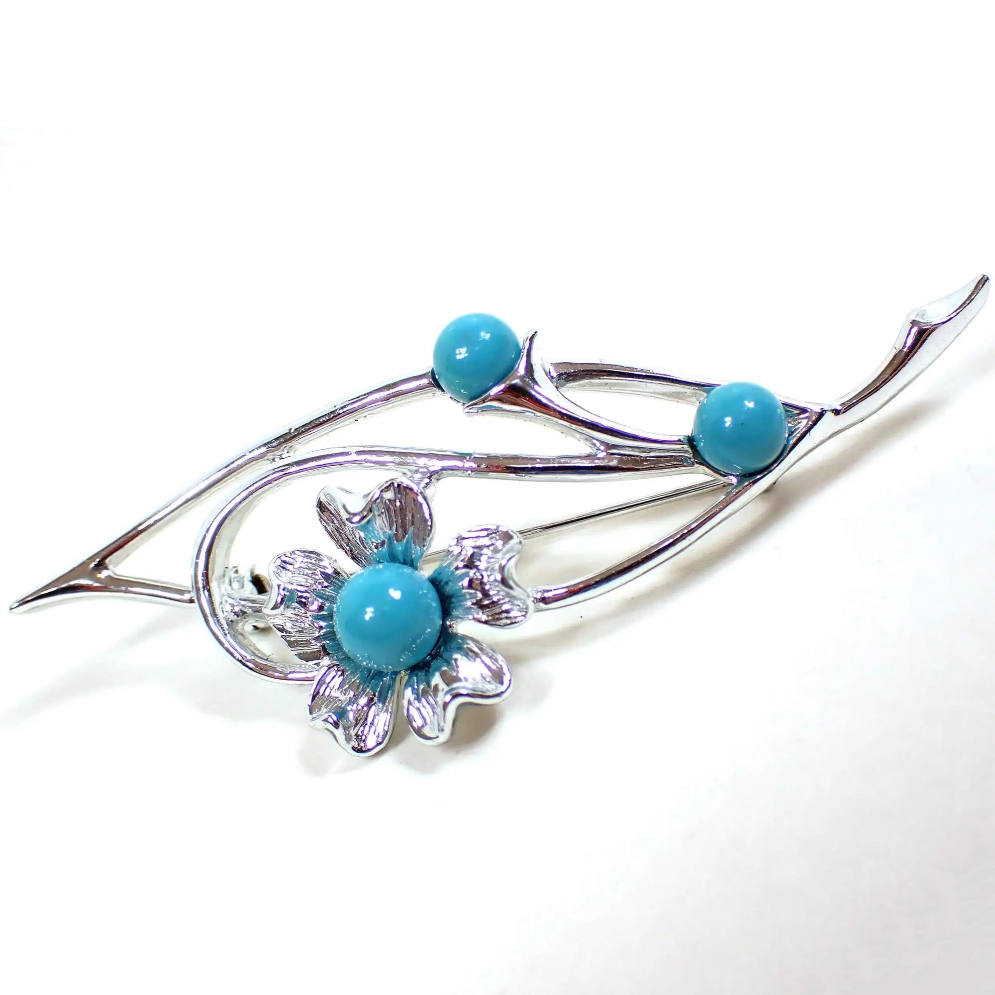 Angled front view of the Mid Century vintage Sarah Coventry floral brooch pin. The metal is silver tone in color. There is an angled open leaf like shape with a curled area and a flower at the end. There are three turquoise blue color cabs on the brooch, including one in the middle of the flower.