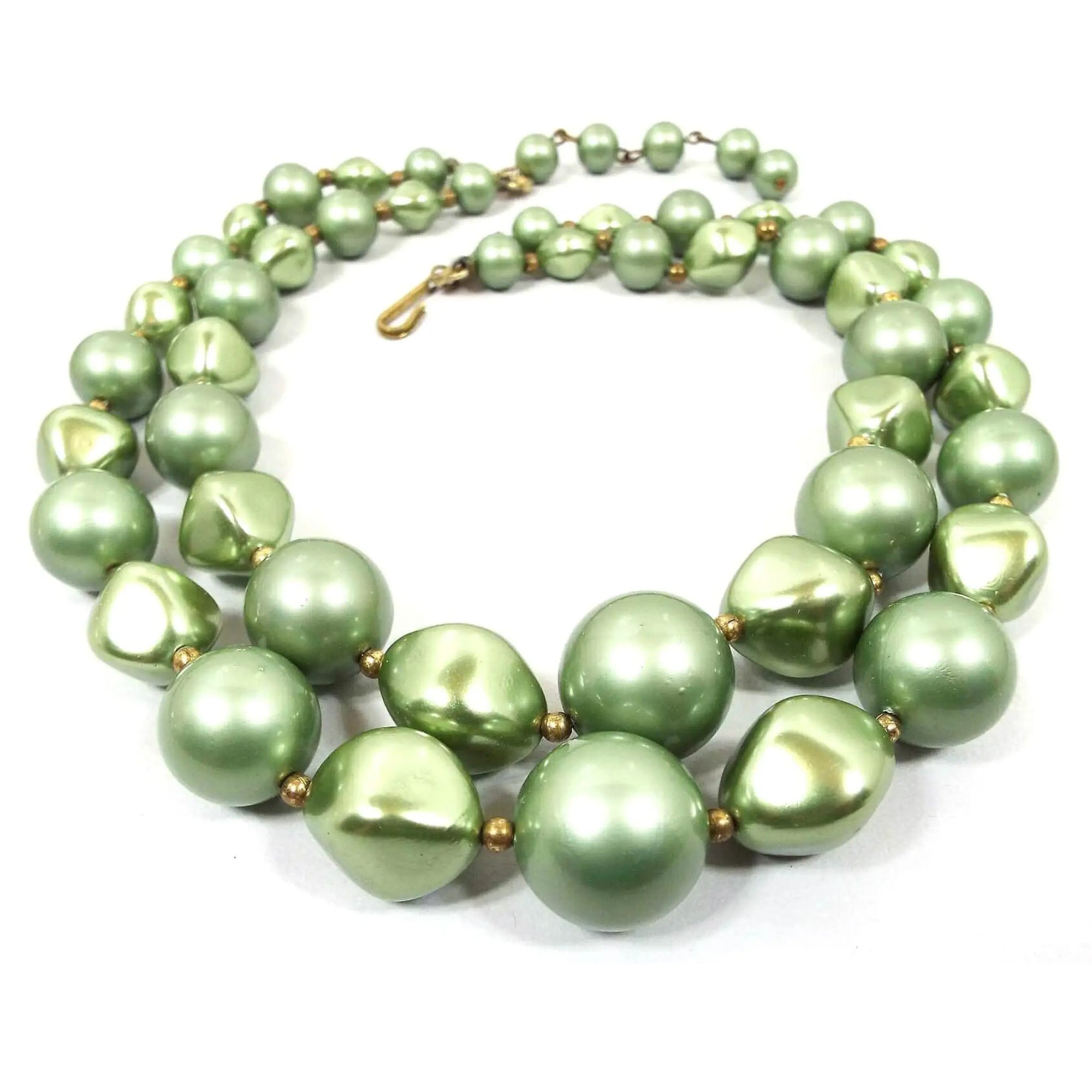 Top view of the Mid Century vintage multi strand beaded necklace. The necklace has two strands of beads. The beads alternate between round and angled oval and are sage green in color. There is a gold tone hook clasp at the end.
