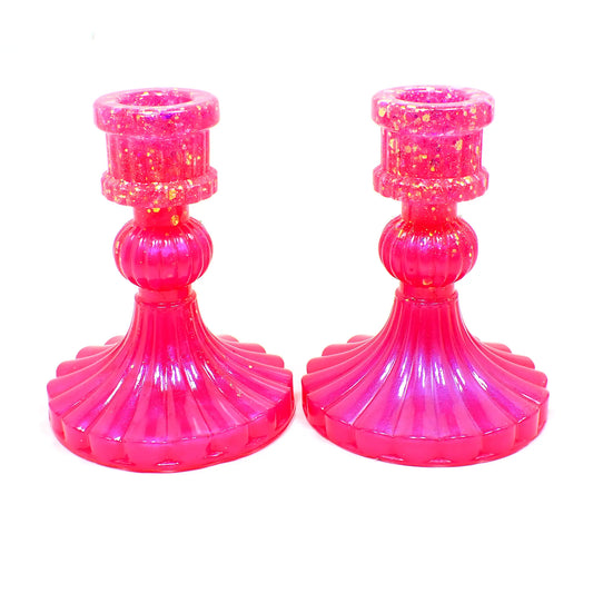 Side view of the vintage style handmade resin candlestick holders. They have a round cylinder style shape at the top, a corrugated round middle, and a flared out bottom with a corrugated ripple design. The resin is bright pearly pink in color and there is iridescent chunky glitter towards the top of the holders.