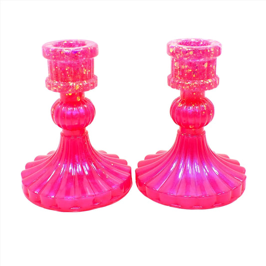 Side view of the vintage style handmade resin candlestick holders. They have a round cylinder style shape at the top, a corrugated round middle, and a flared out bottom with a corrugated ripple design. The resin is bright pearly pink in color and there is iridescent chunky glitter towards the top of the holders.