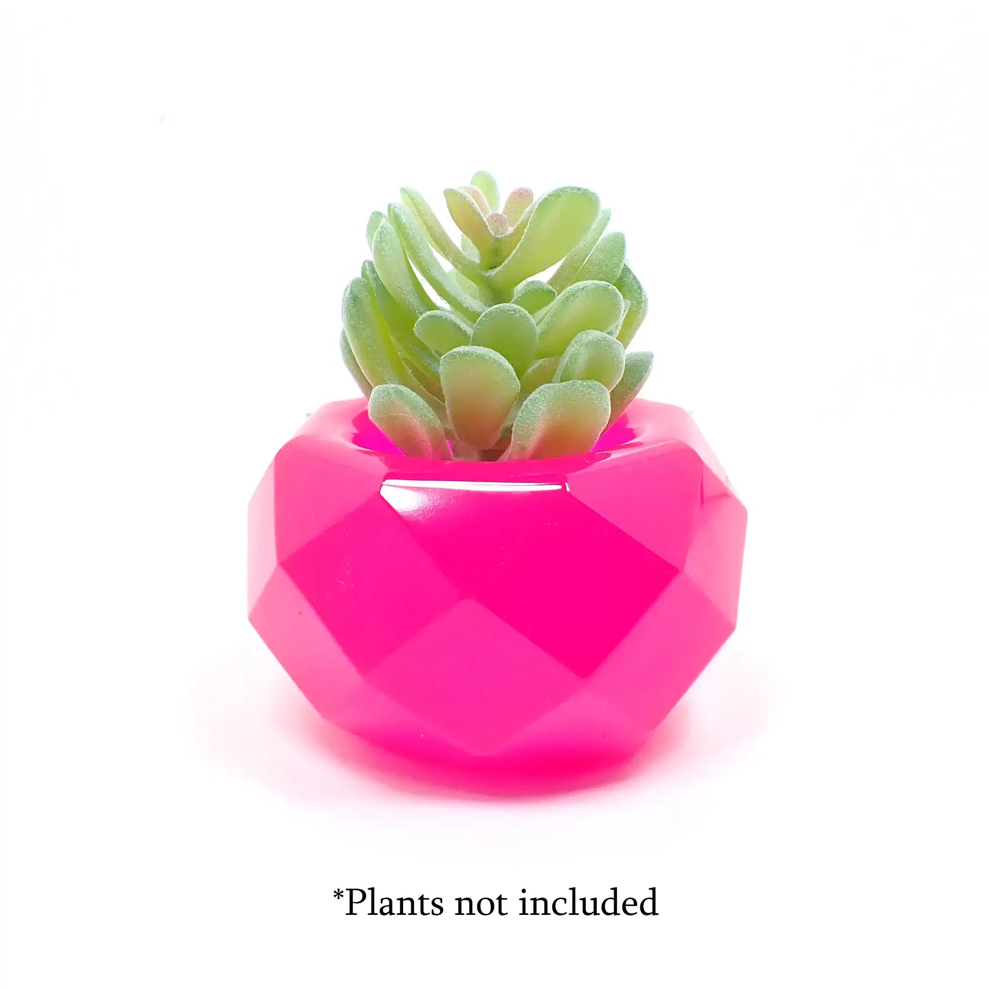 Side view of the small geometric handmade resin succulent pot showing how a small plant fits in the top. The resin is bright neon pink and there is a small fake succulent sitting on top. At the bottom of the photo are the words "Plants not included."