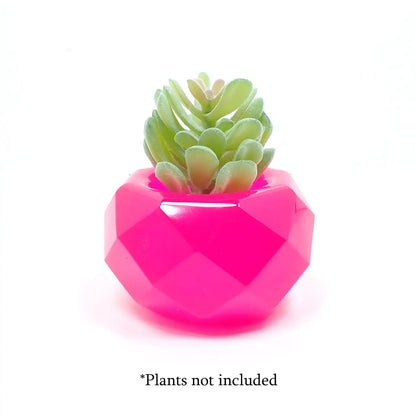 Side view of the small geometric handmade resin succulent pot showing how a small plant fits in the top. The resin is bright neon pink and there is a small fake succulent sitting on top. At the bottom of the photo are the words "Plants not included."