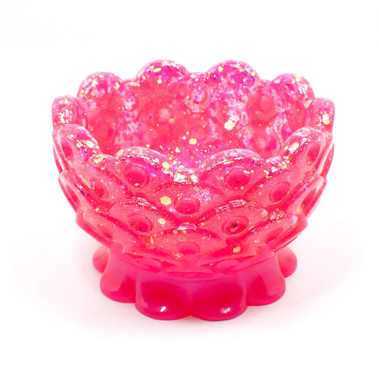 Angled side view of the small handmade resin footed bowl trinket dish. The resin is bright pearly pink in color and has chunky iridescent glitter on the bowl portion of the piece. The top edge and bottom foot area have a scalloped edge. The bowl has a fish scale like design with an indented dot inside each scale area.