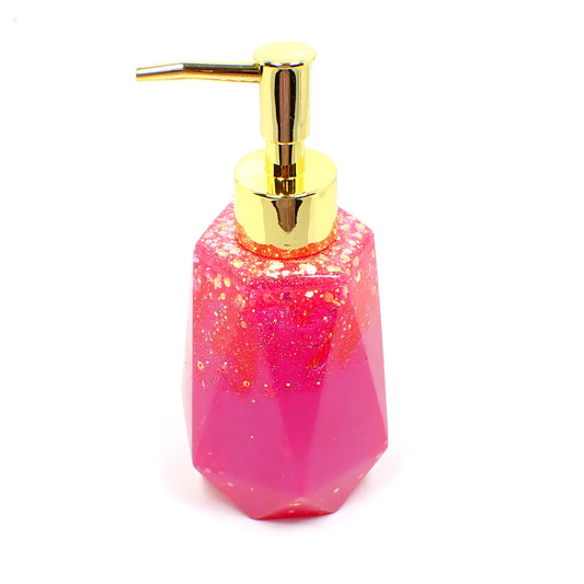 Side view of the handmade resin soap dispenser. In this photo it has a gold tone color pump on it. It is bright pearly pink in color with chunky iridescent glitter mainly on the upper portion of the piece. It has a faceted shape that is narrower at the top and flared at the bottom.