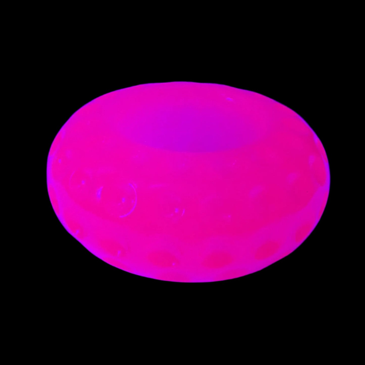 Photo showing the handmade neon pink resin round small pot under a UV light. The pot is a short round saucer shape and has indented circles all the way around it. In this photo it is fluorescing bright pink under the UV light. 