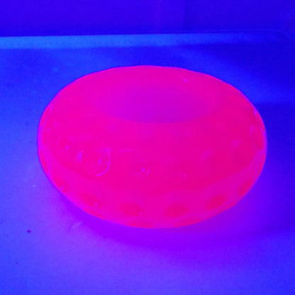 Photo showing the handmade neon pink resin round small pot under a UV light. The pot is a short round saucer shape and has indented circles all the way around it. In this photo it is fluorescing bright pink under the UV light. 