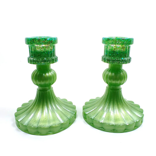 Side view of the vintage style handmade resin candlestick holders. They have a round cylinder style shape at the top, a corrugated round middle, and a flared out bottom with a corrugated ripple design. The resin is pearly lime green in color and there is iridescent chunky glitter at the top.