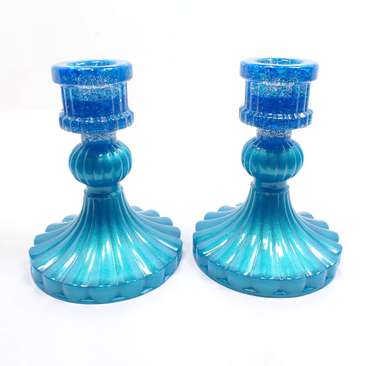 Side view of the vintage style handmade resin candlestick holders. They have a round cylinder style shape at the top, a corrugated round middle, and a flared out bottom with a corrugated ripple design. The resin is pearly aqua blue in color and there is iridescent chunky glitter at the top.