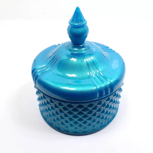 Side view of the pearly aqua blue trinket candy dish. The bottom part has a textured bumpy diamond shaped design all the way around it. The piece itself has a rounded shape. The lid is smooth with some indented lines going down the curves as it tapers at the top. The very top of the lid has a tapered cone shape to grab onto so you can lift the lid off the trinket box.