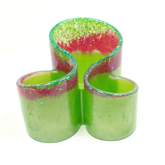 Front view of the handmade resin makeup brush holder. It has pearly lime green resin on the majority of the piece with a strip of iridescent pink glitter towards the top and iridescent green glitter at the very top. There are three round areas to hold the brushes with the back having the tallest side.