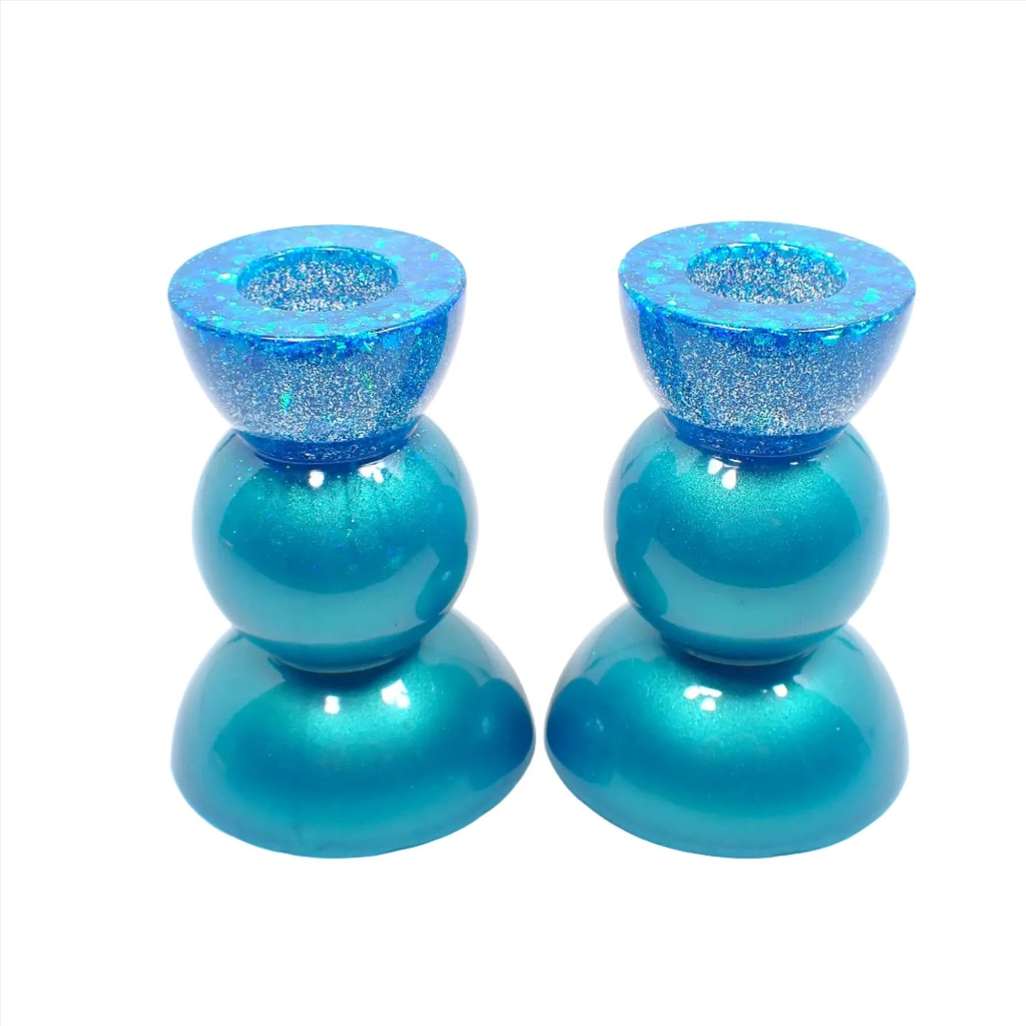 Side view of the handmade resin rounded geometric candlestick holders. They are pearly aqua blue in color with chunky iridescent glitter at the top. They are shaped with a semi circle at the top and bottom with a sphere shape in between. 