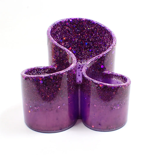Front view of the handmade resin makeup brush holder. It is a bright pearly lilac purple in color with chunky iridescent purple glitter on top. It has three open areas with a curved design to hold the brushes.