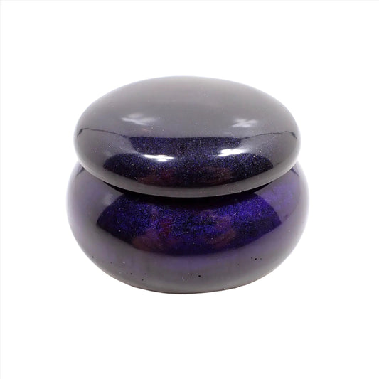 Side view of the small handmade round trinket box jar with lid. It is dark in color with hints of dark pearly purple and blue depending on how the light hits it.