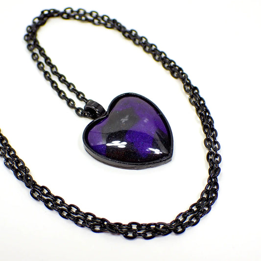 Enlarged view of the handmade Goth heart pendant necklace. The metal chain and pendant setting is black coated base metal in color. The pendant is heart shaped and has a marbled area of pearly black and bluish purple resin.