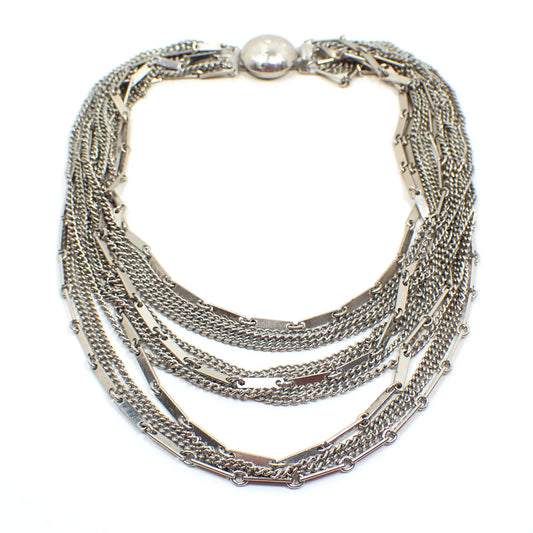 Top view of the Mid Century vintage multi strand necklace. There are thirteen thinner strands of chain. Some are curb link and others are flat bar link chain. There is a round box clasp at the end. The metal is silver tone in color.