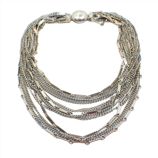 Top view of the Mid Century vintage multi strand necklace. There are thirteen thinner strands of chain. Some are curb link and others are flat bar link chain. There is a round box clasp at the end. The metal is silver tone in color.