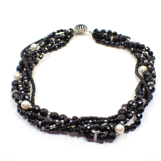 Angled view of the Mid Century vintage beaded multi strand necklace.  There are five strands that mostly consist of black faceted glass beads of different sizes. There are also some small silver tone beads, plastic faux pearl beads, and rhinestone disc beads mixed throughout. The strands are twisted along the necklace and there is a round box clasp at the end with a black glass top.
