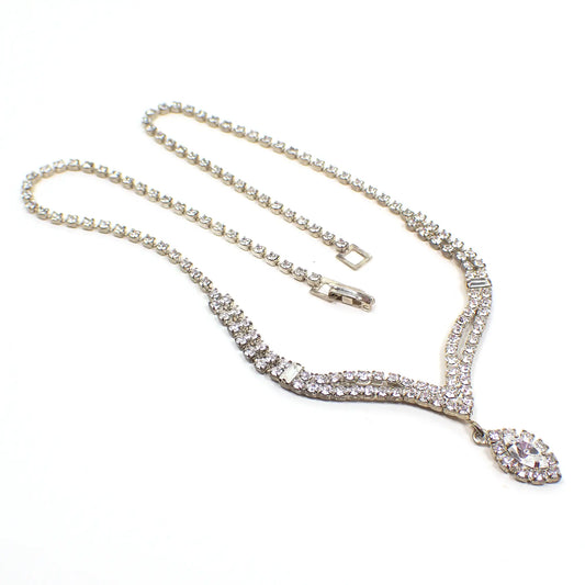 Angled view of the 1980's rhinestone necklace with dangling marquis drop. The metal is silver tone in color. There is a snap lock clasp at the end. The necklace has a single strand of clear round rhinestones that go down to a double strand with a single baguette rhinestone on each side. The double strand part comes down to a curvy V shape and has a dangling marquis shaped drop at the bottom of it that is surrounded with clear round rhinestones. 
