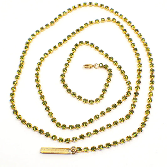 Top view of the retro vintage Liz Claiborne rhinestone necklace. The metal is gold tone in color. The necklace has a long single strand of small round peridot green color rhinestones all the way down the necklace. There is a lobster claw clasp on one side and a hang tag at the other end. 