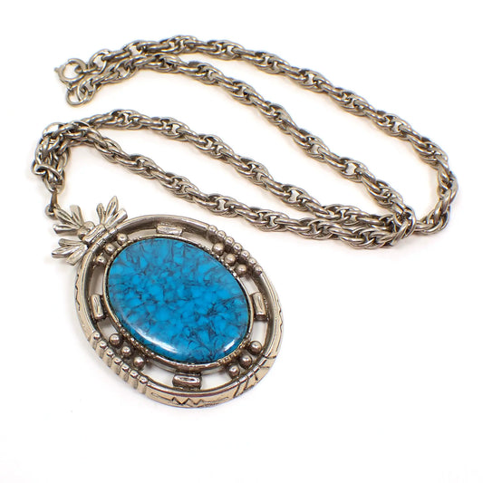 Top view of the retro vintage pendant necklace by Art. The metal is antiqued silver tone in color. The rope chain has a spring ring clasp at the end and a large oval pendant on the bottom. The pendant has a blue and gray marbled plastic cab made to look like turquoise.