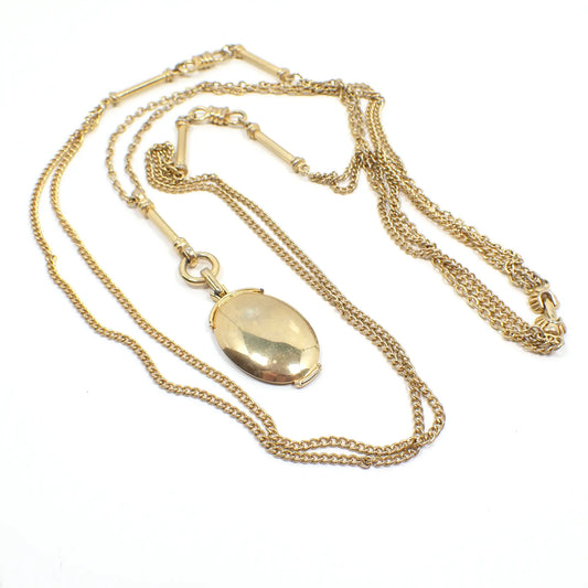 Top view of the Mid Century vintage multi strand chain necklace with pendant. The metal is gold tone in color. There are three strands of chain. Two are curb link and the last is curb link with fancy bar and figure 8 links on it. The pendant hangs from a bar and ring bail and is oval in shape.