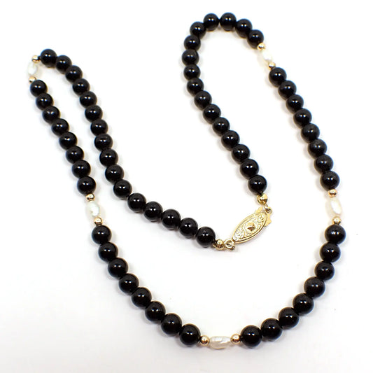 Top view of the retro vintage beaded necklace. There are round black glass beads on the majority of the necklace. There are five small rice shaped glass faux pearl beads spread throughout. At the end is a pearl necklace style clasp that is oval shaped with a hook that snaps inside of it.