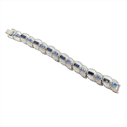 Angled view of the 1950's Mid Century vintage BSK rhinestone link bracelet. The metal is textured matte silver tone in color.  There are light blue round rhinestones links with dark blue baguette rhinestone links in between. There is a snap lock clasp on the end.