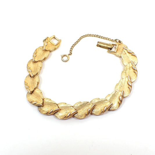 Top view of the Mid Century vintage link bracelet. The metal is textured and gold tone in color. Each link is shaped like a leaf with a curled edge. There is a snap lock clasp and safety chain at the end. 