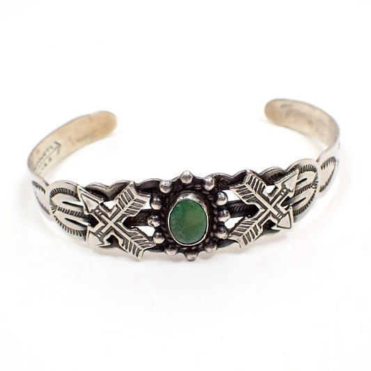 Angled view of the Fred Harvey Era Arrow Novelty coin silver cuff bracelet. It is antiqued silver tone in color. The middle has a small oval green turquoise cab. There is a crossed arrow design on each side and stamped design on the rest of the cuff. Part of the Maker's mark can be seen in the photo.