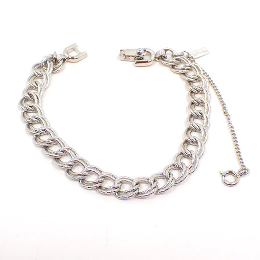 Top view of the Mid Century vintage Monet chain bracelet. It is silver tone in color and has a double link curb chain design. There is a snap lock clasp at the end and a thin safety chain that has a spring ring clasp. 