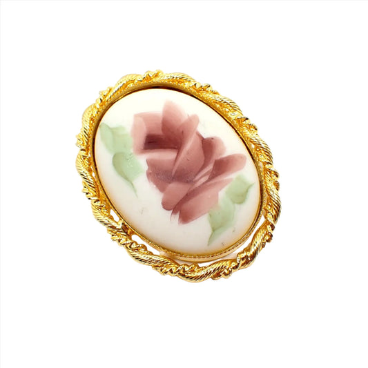 Front view of the retro vintage flower brooch pendant. It is oval in shape with a gold tone metal frame. There is a white porcelain cab in the middle that has a pink flower and green leaves painted on it.