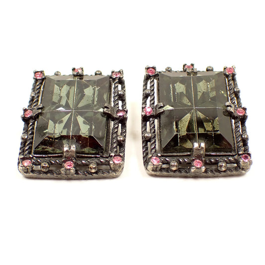 Front view of the Mid Century vintage Sarah Coventry Midnight Magic vintage clip on earrings. They are large rectangle shaped with large rectangle smoky gray rhinestones in the middle and small pink rhinestones around the end. The metal is matte black painted.