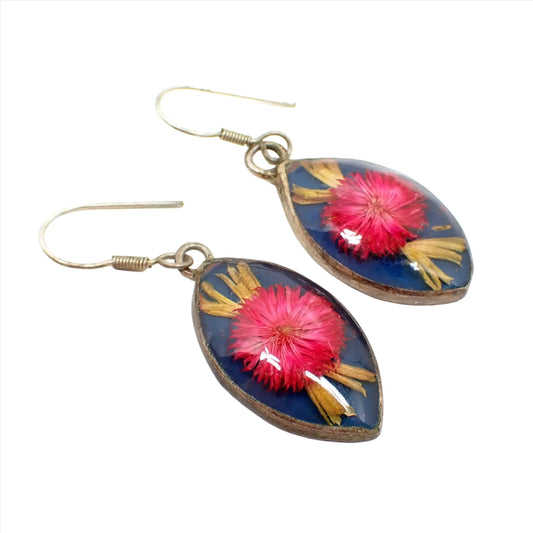 Angled view of the retro vintage Mexican flower and resin earrings. The metal has a darkened patina for a silver and gray color. There are hook earwires at the top. The bottom drops are marquis shaped and have small round pink flowers with light green leaves coming from the top and bottom and a blue background. There is clear resin over the top.