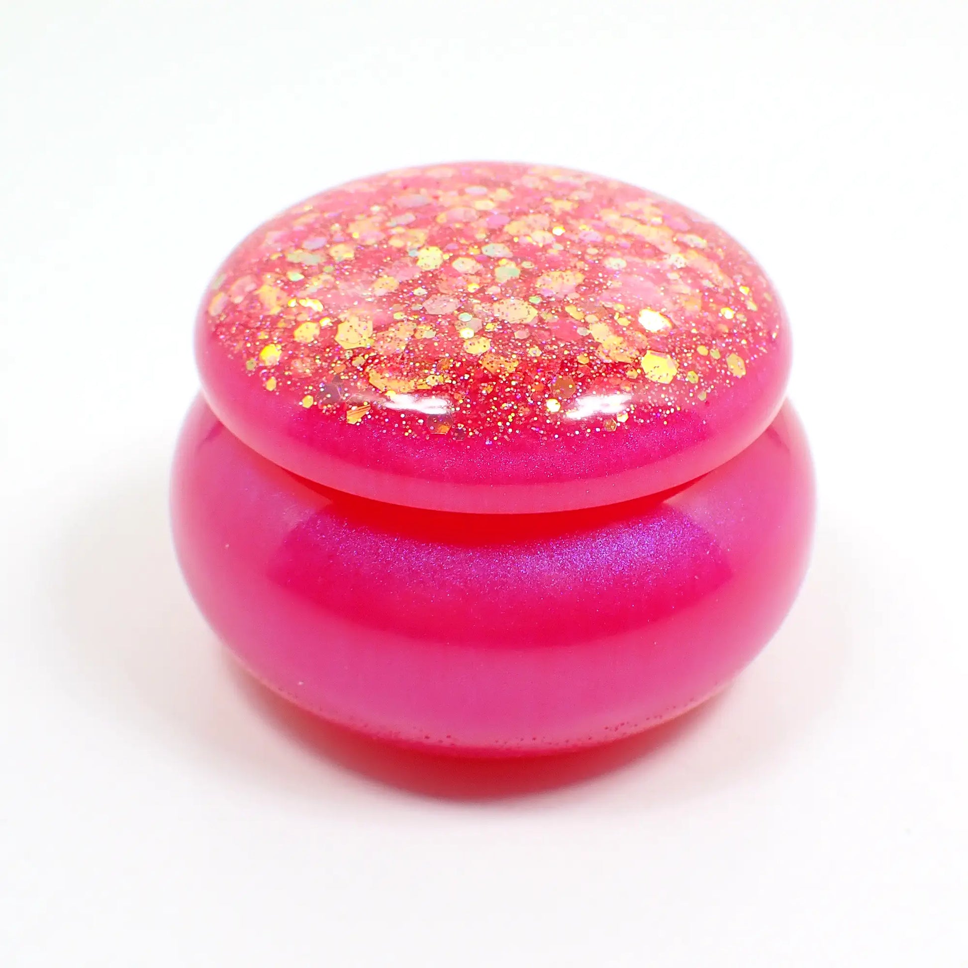 Side view of the small handmade resin trinket jar. It has a rounded shape. The bottom has pearly pink resin and the lid has chunky iridescent pink glitter.