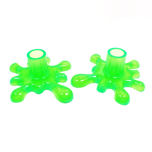 Angled view of the handmade resin larger sized splat style candlestick holders. They are bright neon green in color. The top part that holds the candle is round tube shaped. The bottom has a splat drip style design that is asymmetrical and has rounded ends.