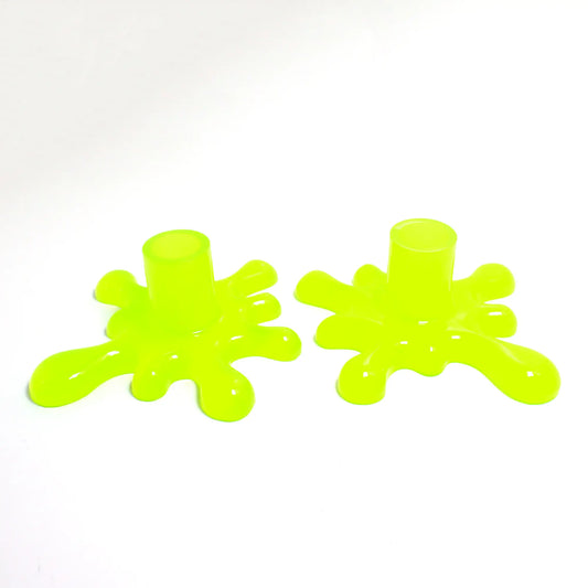 Angled view of the handmade resin larger sized splat style candlestick holders. They are bright neon yellow in color with a tint of bright green. The top part that holds the candle is round tube shaped. The bottom has a splat drip style design that is asymmetrical and has rounded ends.