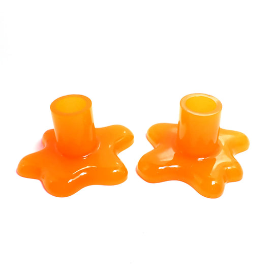 Photo of the handmade resin splat style candlestick holders. They are neon orange in color. The top part that holds the candle is round tube shaped. The bottom has a splat drip style design that is asymmetrical and has rounded ends.