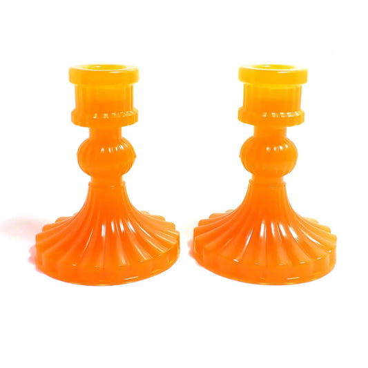 Side view of the vintage style handmade resin candlestick holders. They have a round cylinder style shape at the top, a corrugated round middle, and a flared out bottom with a corrugated ripple design. The resin is neon orange in color. 