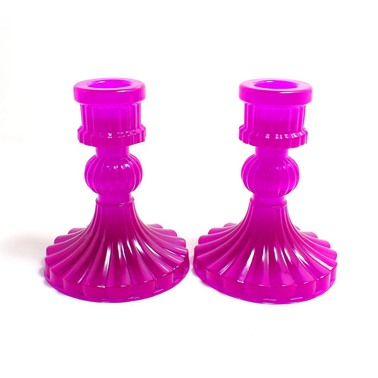 Side view of the vintage style handmade resin candlestick holders. They have a round cylinder style shape at the top, a corrugated round middle, and a flared out bottom with a corrugated ripple design. The resin is neon purple in color. 
