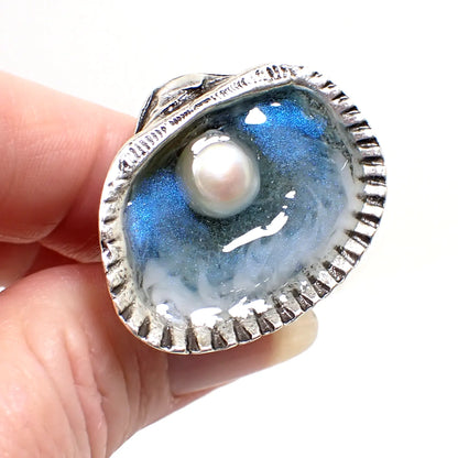 Angled view of the handmade seashell and cultured pearl ring. The metal is antiqued silver tone in color. It is shaped like a half seashell on top. There is a real cultured pearl surrounded by iridescent blue and clear resin and wisps of white resin for waves. There is beach sand at the bottom of the ring.