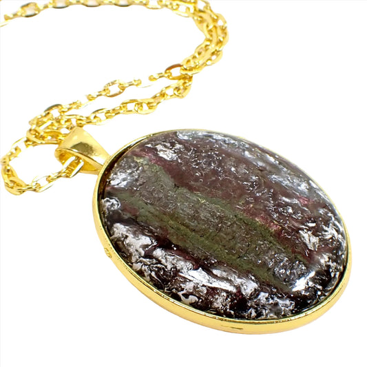 Enlarged view of the handmade pendant with vintage abalone and resin cab. The metal is gold tone in color. There is a cable chain running through the pendant bail. The pendant is a large oval with a slightly domed cabochon that has iridescent shades of green, brown, and red with areas of frosty white in the top layers of resin.