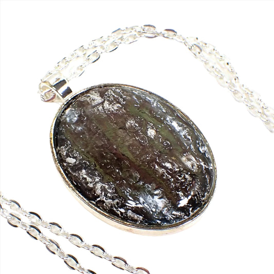 Enlarged view of the handmade pendant with vintage abalone and resin cab. The metal is silver tone in color. There is a cable chain running through the pendant bail. The pendant is a large oval with a slightly domed cabochon that has iridescent shades of green, brown, and red with areas of frosty white in the top layers of resin.