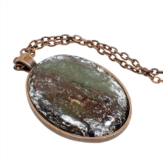 Enlarged view of the handmade pendant with vintage abalone and resin cab. The metal is antiqued copper in color. There is a cable chain running through the pendant bail. The pendant is a large oval with a slightly domed cabochon that has iridescent shades of green, brown, and red with areas of frosty white in the top layers of resin.