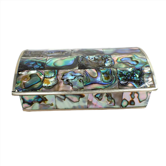 Angled view of the 1970's Mexican Alpaca and abalone shell jewelry box. It is rectangle shaped with a curved top. The outside is covered with rectangle pieces of iridescent multi color abalone shell. The metal trim is silver tone in color.