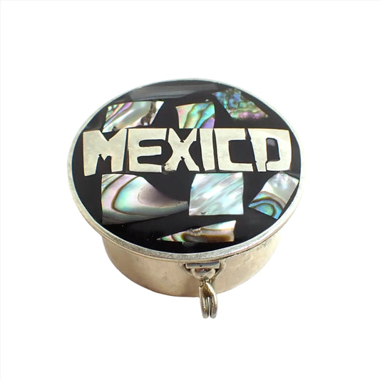 Angled view of the small vintage Alpaca pillbox. It is round with silver tone color metal. The top is black enameled with "Mexico" in the silver color metal. There are inlaid pieces of iridescent multi color abalone shell around it. The clasp on the side can be seen in the photo.