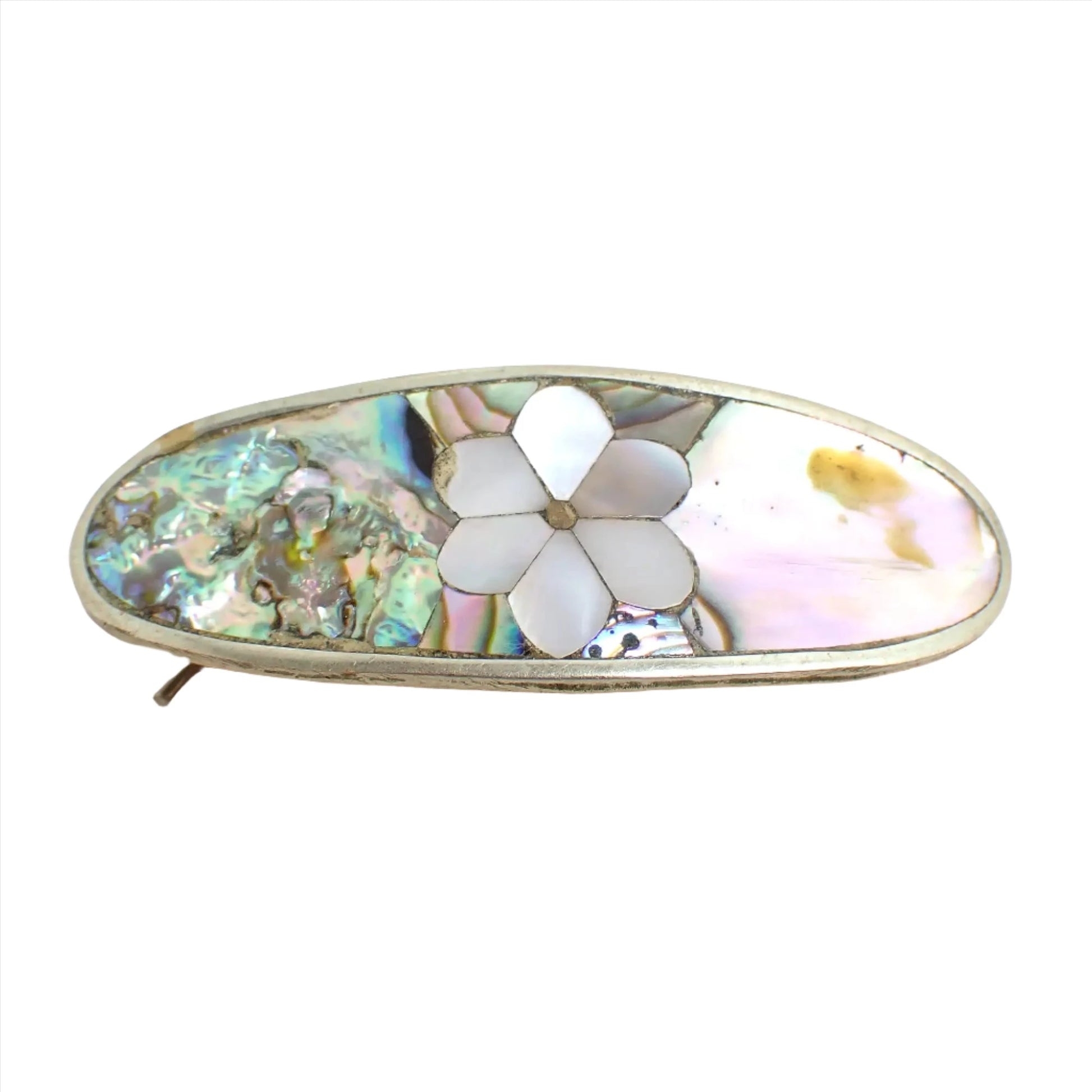 Top view of the 1980's Alpaca Mexico small vintage hair clip barrette. It is oval shaped with silver tone color metal. In the middle is inlaid pearly white mother of pearl shell in a flower design. The rest of the piece has iridescent multi color inlaid abalone shell. 