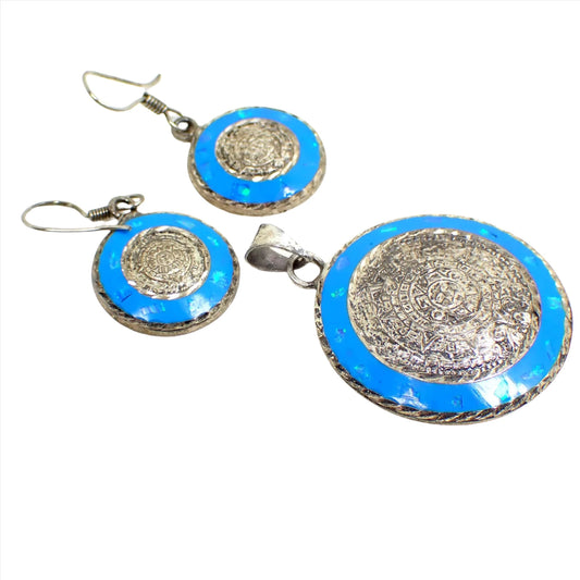 Top view of the 1980's vintage Mexican Jewelry Set.  There is a pair of hook pierced earrings and a pendant. All pieces have round drops with silver tone color metal. The edges have blue enamel with pieces of iridescent lab created opal. The middles have a depiction of the Mayan Calendar. The pendant is by itself and has no chain.