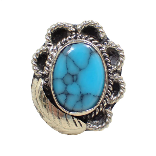 Front view of the Mexican vintage faux turquoise ring. The ring is silver tone in color with a scalloped design around the edge and a feather shape on one corner of the ring. The front cab is made of resin and is blue in color with gray and black lines to make it look like turquoise. 